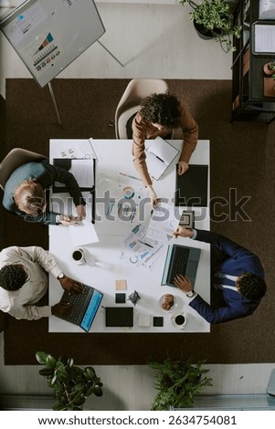 Tabletop view of three professionals working on statistical charts on desk surrounded by plants and equipment. Business meeting involving laptop and documents