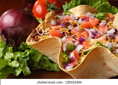 Taco salad in freshly baked flour tortilla bowl (seasoned ground beef, lettuce, onions, tomatoes, and shredded cheese) with tomato, onion, and leaf lettuce in background.  Macro with shallow dof.