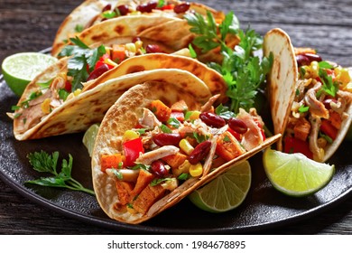 tacos of flour tortilla shells filling with grilled chicken meat, corn, roasted sweet potatoes cubes, red pepper and parsley served on a black plate, close-up