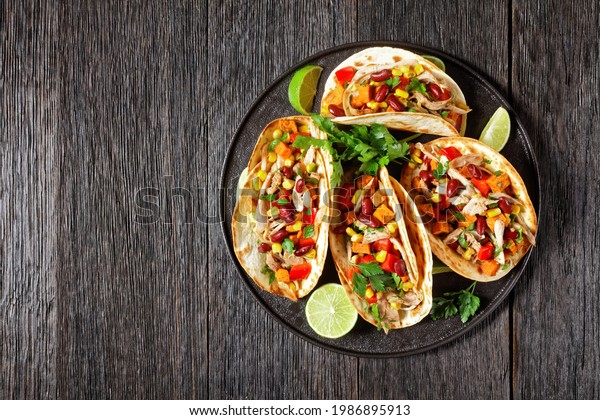 tacos of flour tortilla\
shells filling with grilled chicken meat, corn, roasted sweet\
potatoes cubes, red pepper and parsley served on a black plate,\
flat lay, free space