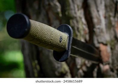 A tactical knife is embedded in a tree trunk. The knife features a black blade and a textured green handle. The blade is sharp and extends outward from the tree, while the handle appears sturdy.