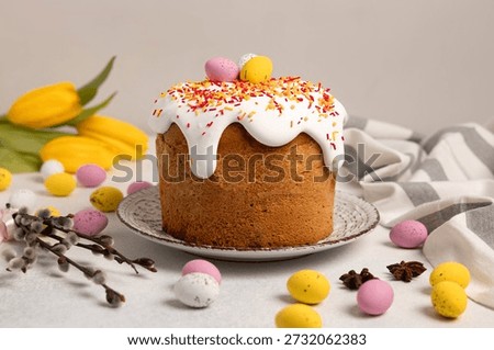 Taditional Easter cake decorated with white icing, colorful sprinkles, and pastel eggs and served on a beautiful plate. Pussy willow branches, yellow tulips and chocolate Easter eggs lying nearby.