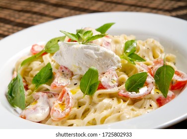 Tagliatelle italian pasta, topped with white goat cheese, basil leaves and cherry tomatoes, served on a plate.