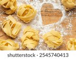 Tagliatelle pasta nests are arranged on a wooden board sprinkled with flour, forming a heart shape