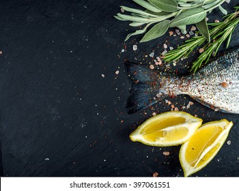 Tail of fresh raw Dorado or sea bream fish on black slate stone board with spices, herbs, lemon and salt. Top view, copy space