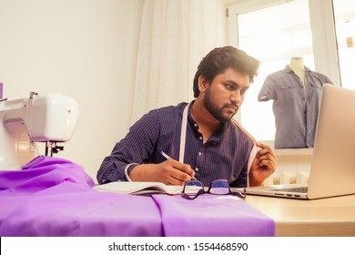 Tailor Asian Man Designer Sewing Clothes On Sewing Machine Next To Laptop Studio Workplace.