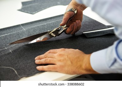 Tailor cutting out the marked pattern on dark fabric with large scissors on the workbench in his shop, close up view of his hands