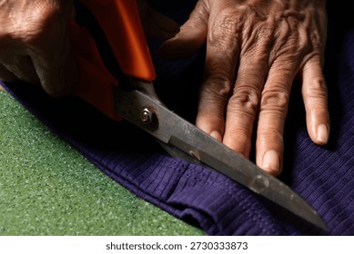 A tailor using orange-handled scissors to cut purple ribbed fabric, focusing on manual dexterity and traditional craftsmanship skills