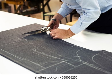 Tailor working in his shop cutting a roll of dark fabric on which he has marked out the pattern of the garment he is making with tailors chalk, close up view