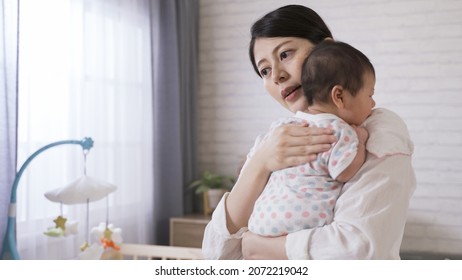 Taiwanese new mother is standing up from the bed and holding her baby properly while burping in the bedchamber at home.