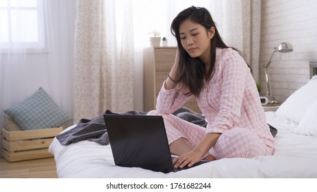 Taiwanese Young Woman Is Teleworking With Laptop In Pajamas During Virus Lockdown. Female Blogger Sitting On Bed Looking Thoughtful While Writing Articles On Computer. Work From Home Concept