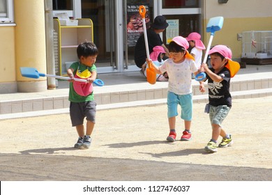 TAKAYAMA, JAPAN - June 4, 2018. Little Japanese Boys Are Playing Together With Plastic Scoops In Outdoor Playground Of Kindergarten, While Parents Are Working. Father Says Goodbye To Daughter.