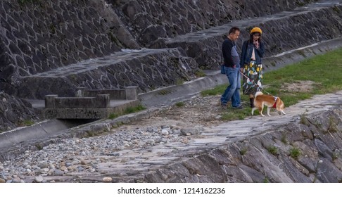 TAKAYAMA, JAPAN - OCTOBER 23RD, 2018. Japanese Couple With Their Pet Dog Walking Along Miyagawa River Banks At Dusk.