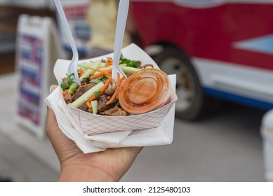 Take away paper food tray of chicken teriyaki with chili sauce in deli containers, napkins, fork and spoon with blurry food truck background. Togo grilled meat with allumette cut cucumber carrot slice