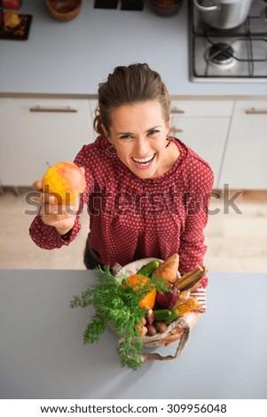 Take a bite, I promise it is delicious... An elegant woman is laughing and teasing in a kitchen, holding up colourful apple and cradling a burlap sac of fresh fall vegetables bought at the market.