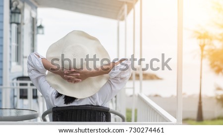 Take it easy lifestyle of carefree young woman wearing summer hat relaxing happily sitting on porch at beach-house on beach front enjoying healthy living life quality for holiday vacation concept