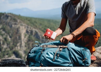 Take a first-aid kit in a backpack on a trip, a hand holds a first aid kit against the background of mountains, equipment in an extreme hike, a white cross