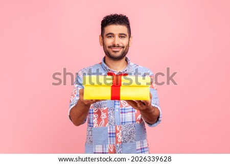 Take it! Young adult happy man wearing blue casual style shirt giving gift box to camera with excited smile, greeting on holiday and sharing present. Indoor studio shirt isolated on pink background.