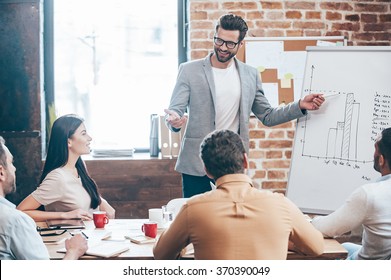 Take a look at our results! Handsome young man in glasses standing near whiteboard and pointing on the chart while his coworkers listening and sitting at the table  - Powered by Shutterstock