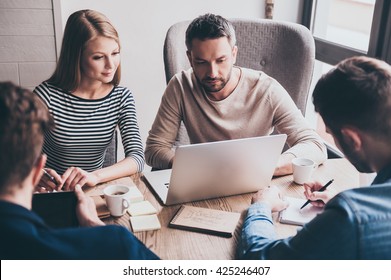 Take A Look At This Chart! Young Handsome Man Using Laptop While Sitting At The Office Table On Business Meeting With His Coworkers