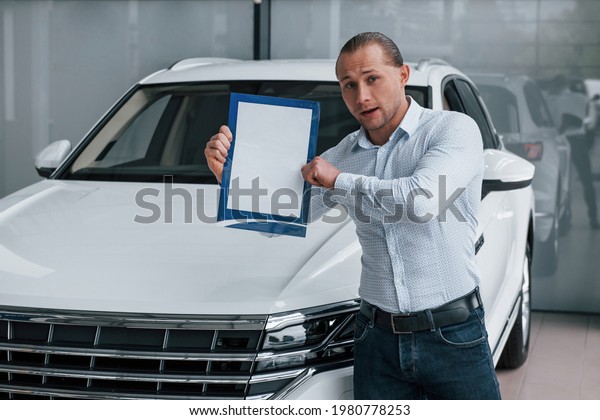 Take a look at this.\
Manager stands in front of modern white car with paper and\
documents in hands.