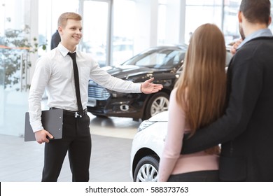 Take a look at this one. Handsome young cheerful salesman smiling showing cars at the dealership to his clients couple buying a car family relationship service advice helpful professionalism lifestyle