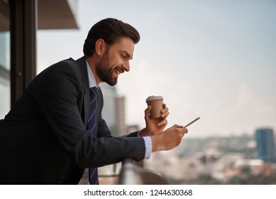 Take A Pause. Waist Up Portrait Of Smiling Businessman Reading Massages On Smartphone While Having Coffee Break On Office Balcony