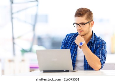 Take Your Time. Pleasant Handsome Young Guy Sitting At The Table And Working On The Computer While Concentrating On His Work