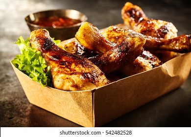 Takeaway box of crispy grilled chicken legs with a spicy marinade served with fresh lettuce and a hot chili dipping sauce, close up view