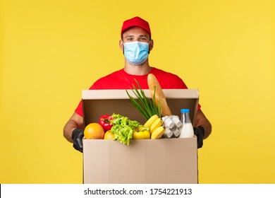 Takeaway Delivery, Covid-19 Quarantine And Groceries Concept. Happy Smiling Courier In Red Uniform And Medical Mask Bring Grocery Box To Customer House, Buying From Home