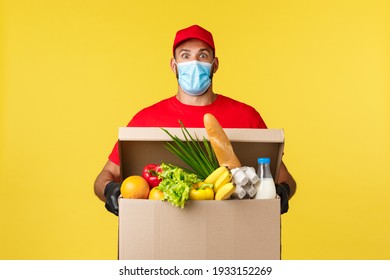 Takeaway Delivery, Covid-19 Quarantine And Groceries Concept. Surprised Courier In Medical Mask, Uniform Red Cap And T-shirt, Staring Shocked, Bring Food Order Box To Customer Home, Yellow Background