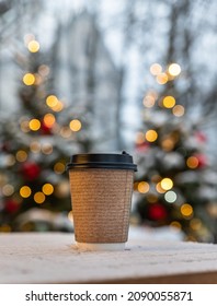 Takeaway Disposable Cup For Hot Drink On Wooden Table. Street Coffee Shop.Christmas Winter Atmosphere.Coffee, Tea Or Mulled Wine To Go. Empty Space For Text, Mockup. Blurred Background With Garland.