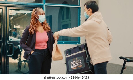 Takeaway employee with face mask giving food order to african american woman at front door, delivery service during covid 19 pandemic. Courier delivering fastfood restaurant package. - Powered by Shutterstock