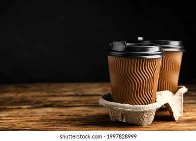 Takeaway paper coffee cups in cardboard holder on wooden table against black background, space for text