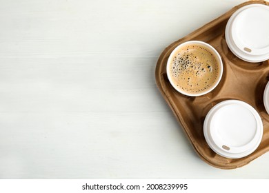 Takeaway paper coffee cups in cardboard holder on white wooden table, top view. Space for text