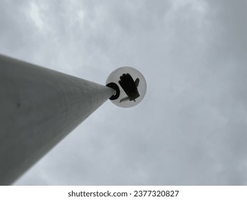 Taken from below, the image presents a distinctive perspective. A broken light bulb hangs at the top of an intact plastic lamp pole, casting a hand-shaped shadow through a football glove. 