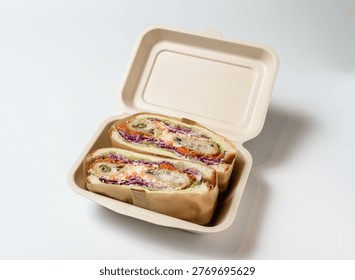 A takeout container made of biodegradable material holds two halves of a sandwich with vibrant purple cabbage, green lettuce, orange carrots, and sliced jalapeños on a white surface