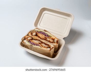 A take-out container made of biodegradable material holds two halves of a sandwich, filled with chicken, purple cabbage, and avocado, on a clean white surface with subtle shadows