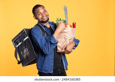 Takeout service employee carrying bag full of fresh groceries, bringing organic produce order to customers. African american express courier holding local eco friendly fruits and vegetables. - Powered by Shutterstock