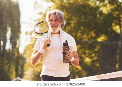 Taking break and drinking water. Senior stylish man in white shirt and black sportive shorts on tennis court. - Powered by Shutterstock