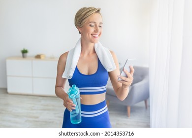 Taking Break. Happy blonde woman with white towel on shoulders holding bottle of water and using smartphone standing in living room at home, resting, free copy space. Sports application concept - Powered by Shutterstock