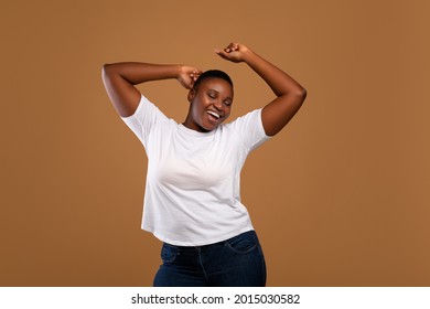 Taking Break. Overjoyed millennial plus-size black woman wearing white t-shirt dancing with closed eyes, having fun moving and listening to music, relaxing isolated over dark brown studio background