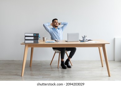 Taking Break. Smiling young Arab man relaxing on chair sitting at table and resting, using pc laptop, happy millennial male leaning back at workplace, enjoying his job, feeling pleased and satisfied