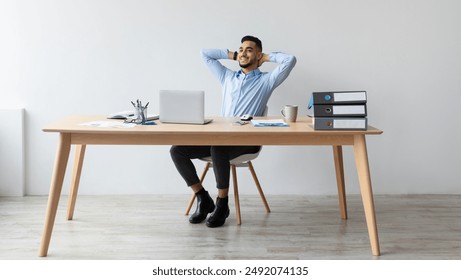 Taking Break. Smiling young Arab man relaxing on chair sitting at table and resting, using pc laptop, happy millennial male leaning back at workplace, enjoying his job, feeling pleased and satisfied - Powered by Shutterstock