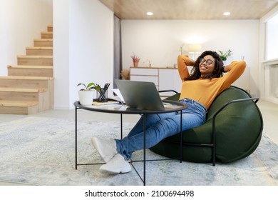 Taking Break. Smiling young black woman in glasses relaxing on bean bag chair sitting at tea table and resting, using pc laptop, happy millennial female leaning back, enjoying job, feeling pleased - Powered by Shutterstock