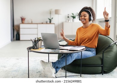 Taking Break From Work. Joyful casual black lady in wireless headphones dancing, having fun and listening to music, sitting on bean bag pouf chair with pc on coffee tea table, free copy space