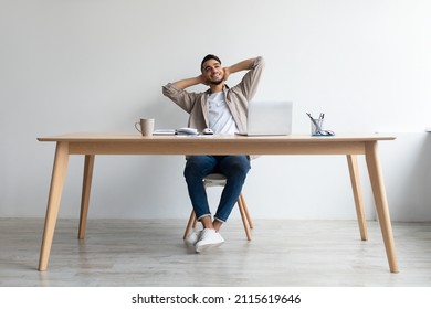 Taking Break From Work. Portrait of smiling young Arab man relaxing on chair sitting at table and using laptop, happy millennial male leaning back at workplace and dreaming, holding hands behind head