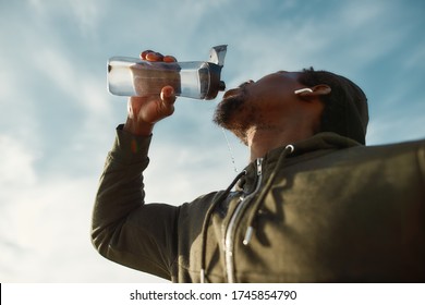 Taking a break. Young african male runner in sportswear drinking fresh water from a bottle while standing against blue sky. Jogging at sunrise, sport, cardio, healthy lifestyle. Refreshing