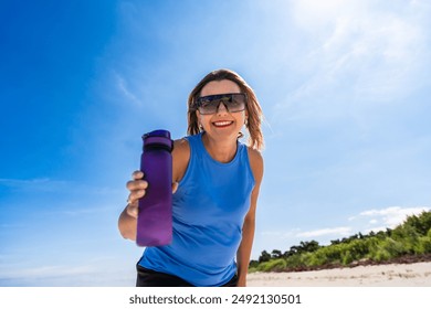 Taking care to drink water in hot summer. Portrait of young modern woman with long blonde hair in blue top and sunglasses giving water bottle on beach by sea on sunny day. Health, energy, joy of life - Powered by Shutterstock