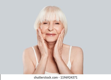 Taking Good Care Of Her Skin. Beautiful Senior Woman Looking At Camera And Smiling While Standing Against Grey Background